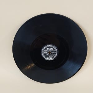 Pode incluir: Um disco de vinil preto com um rótulo circular no centro. O rótulo apresenta texto cirílico e um design circular. O disco está sobre um fundo bege claro. As ranhuras do disco são visíveis.