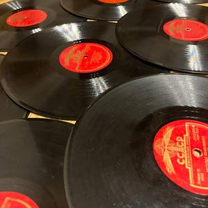 May include: A collection of black vinyl records with red labels. The records are scattered across a surface, showcasing their circular shape and the details of the labels. The labels feature text and designs.