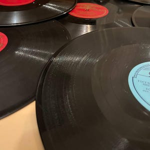 Pode incluir: Uma coleção de discos de vinil pretos com rótulos vermelhos e brancos. Os discos estão dispostos em um padrão ligeiramente sobreposto, mostrando sua forma circular e superfícies reflexivas. A imagem captura a textura e os detalhes do formato de música vintage.