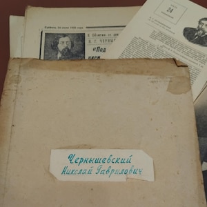May include: A stack of aged papers and documents, including a newspaper with a portrait and the number 24. A label with Cyrillic text is visible, reading "Chernyshevsky Nikolai Gavrilovich."