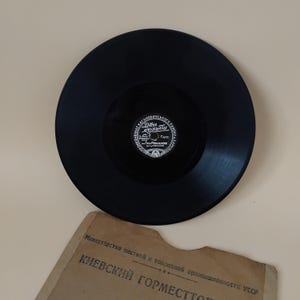 Pode incluir: Um disco de vinil preto com um rótulo branco com texto cirílico. O disco está centrado numa superfície bege clara, com uma capa de papel vintage visível por baixo. A capa tem texto cirílico e parece envelhecida.
