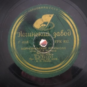 Pode incluir: Close-up de um disco de vinil vintage com um rótulo verde e dourado. O rótulo apresenta texto cirílico, um logótipo e as palavras "Ногинский завод". O disco é preto com um orifício central.