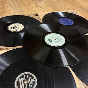 Pode incluir: Uma coleção de discos de vinil vintage pretos, cada um com uma etiqueta circular. As etiquetas variam em cor e design, com texto e ilustrações. Os discos estão dispostos sobre uma superfície de madeira.