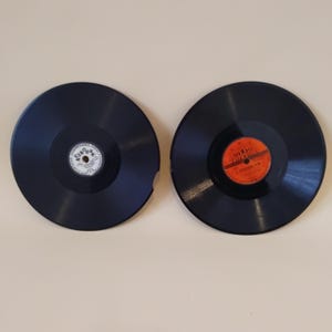 Pode incluir: Dois discos de vinil pretos com rótulos coloridos. Um disco tem um pedaço faltando na borda. Os rótulos apresentam texto em estilo vintage. Os discos estão em um fundo neutro.