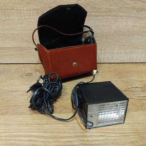 May include: A vintage camera flash unit with a black body and a grid-patterned front. It is connected to a power cord and sits next to a brown leather case with a black interior. The case is open, revealing its interior.