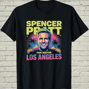May include: Black t-shirt featuring a colorful graphic of Spencer Pratt with the text "Spencer Pratt For Mayor Los Angeles". The design includes palm trees and a cityscape, with text in yellow, pink, and blue.