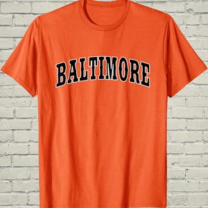 May include: Orange t-shirt with the word "BALTIMORE" in black, outlined in white, across the chest. The shirt has a crew neck and short sleeves. The background is a white brick wall.