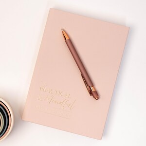 May include: A pale pink daily planner with gold lettering reading "The Practical & Mindful Daily Planner." A matching pen with gold detailing lies on the planner. The planner and pen are on a white surface.