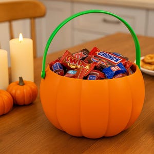 May include: An orange pumpkin-shaped candy basket with a green handle, filled with assorted wrapped sweets. Two small orange pumpkins and two lit candles are on the wooden table. A plate of pastries is in the background.