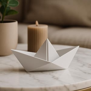 May include: A white paper boat ornament on a marble table. A beige candle and a plant in a pot are in the background. The boat is a decorative item, adding a minimalist touch to the decor.
