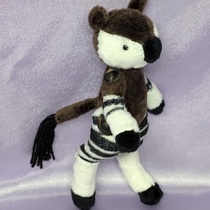 May include: A plush okapi toy with a brown and white body, black and white striped legs, and a black tail. The okapi has button details and is set against a lavender background.