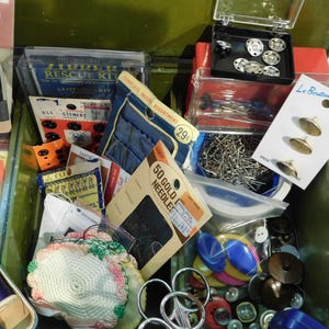 May include: An open green metal sewing box filled with various sewing supplies. The box contains spools of thread, needles, buttons, pins, and other notions. Packaged items include a sewing rescue kit, bias facing, and gold needles.