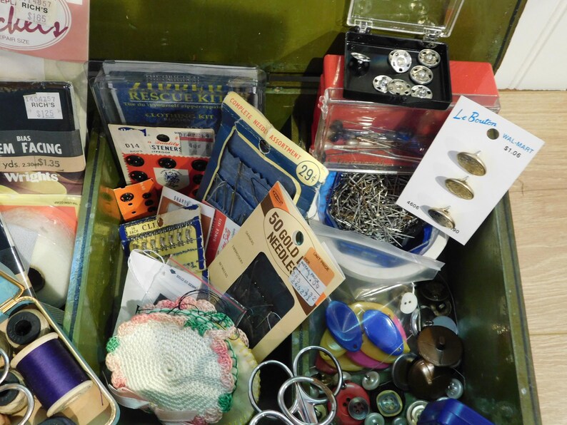 May include: An open green metal sewing box filled with various sewing supplies. The box contains spools of thread, needles, buttons, pins, and other notions. Packaged items include a sewing rescue kit, bias facing, and gold needles.