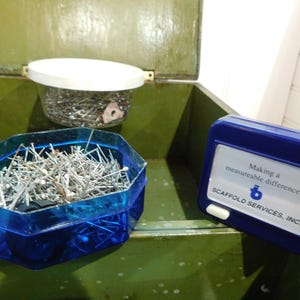 May include: An open green metal box contains two containers of sewing pins and a blue measuring tape. One container is a clear plastic jar with a white lid. The other is a blue, multi-sided container. The measuring tape has text that reads "Making a measureable difference."