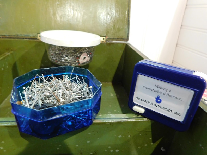 May include: An open green metal box contains two containers of sewing pins and a blue measuring tape. One container is a clear plastic jar with a white lid. The other is a blue, multi-sided container. The measuring tape has text that reads "Making a measureable difference."