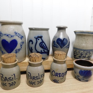 May include: A collection of gray stoneware pottery featuring blue painted designs. The set includes three small jars with cork stoppers labeled Basil, Parsley, and Oregano, a heart-patterned crock, a bird-patterned jar, a heart-patterned bottle, a pitcher, and a small bowl.
