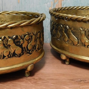 May include: Two oval brass planters featuring a raised relief pattern of rabbits around the exterior. Each planter has a twisted rope-style rim and stands on four small, rounded feet. The brass has a polished, metallic gold-toned finish.