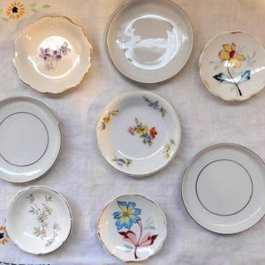 May include: A collection of eight small, vintage porcelain plates. The plates feature floral designs and gold trim. Some plates have scalloped edges, while others are round with a simple gold or silver band.