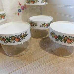 May include: A set of clear glass storage containers with white lids. Each lid has a decorative band featuring colorful illustrations of fruits and vegetables. The containers are arranged on a light-colored wooden surface.