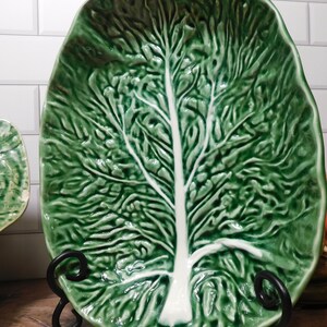 May include: An oval ceramic serving platter shaped like a large green cabbage leaf. The dish features a glossy finish with deep green textured foliage and a prominent, smooth white central vein running vertically through the center of the piece.