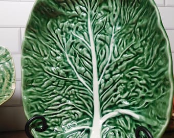 May include: An oval ceramic serving platter shaped like a large green cabbage leaf. The dish features a glossy finish with deep green textured foliage and a prominent, smooth white central vein running vertically through the center of the piece.