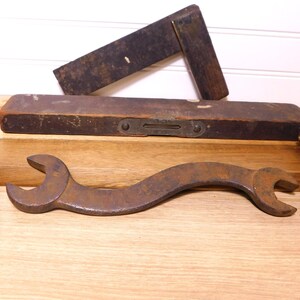 May include: An assortment of vintage carpentry tools, including a rusty wrench, chisels, a level, and a square. The tools are arranged on a wooden surface, showcasing their aged appearance and craftsmanship.