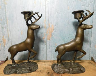 May include: A pair of bronze-toned metal candle holders, each shaped like a standing deer with antlers. The deer are positioned on textured, rocky-looking bases, with a circular candle cup mounted between the antlers of each figure.