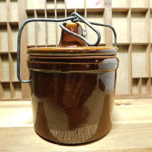 May include: A cylindrical brown glazed ceramic crock with a matching lid and a thick, grey metal wire bail handle. The lid features a central knob, and the entire piece has a glossy finish, reflecting light on its smooth, dark brown surface.