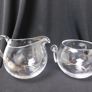 May include: A clear glass creamer and sugar bowl set. The creamer has a curved handle and a spout. The sugar bowl has two small handles. Both pieces are round and sit on a dark surface.