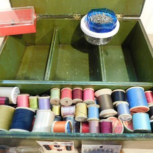 May include: Open green sewing box filled with spools of thread in various colors, including pink, blue, yellow, and green. A red plastic container and a container of pins are also visible. The box is divided into compartments.