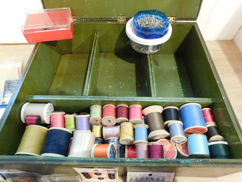 May include: Open green sewing box filled with spools of thread in various colors, including pink, blue, yellow, and green. A red plastic container and a container of pins are also visible. The box is divided into compartments.