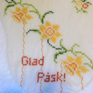 May include: A white fabric runner featuring cross-stitch embroidery of yellow and orange daffodils with green stems and leaves. The text "Glad Påsk!" is stitched in orange thread. The fabric has a scalloped edge with a delicate white trim.