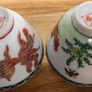 May include: Two antique porcelain bowls, each with a unique hand-painted design. One bowl features a red and white creature, while the other showcases a floral and bird motif. Both have a red stamp on the bottom.