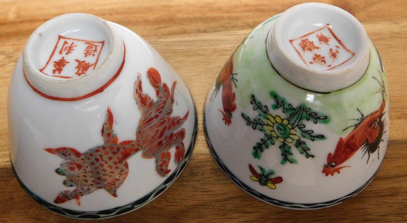 May include: Two antique porcelain bowls, each with a unique hand-painted design. One bowl features a red and white creature, while the other showcases a floral and bird motif. Both have a red stamp on the bottom.