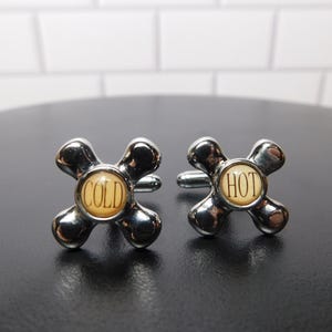 May include: A pair of silver-toned metal cufflinks shaped like vintage cross-handle faucet knobs. The center of each knob features a circular cream-colored inlay with black text, one reading COLD and the other reading HOT, in a classic serif font.