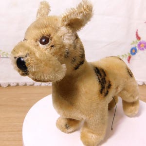 May include: A vintage plush dog toy with a tan body and black stripes. The dog has a black nose, brown eyes, and floppy ears. It is standing on a white surface.