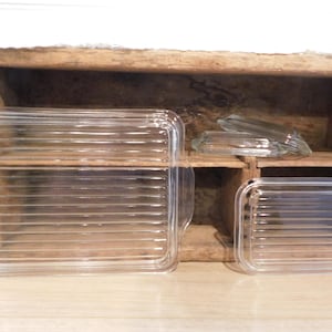 May include: Two rectangular clear glass lids with horizontal ribbed textures and integrated side handles. The larger lid is positioned on the left, while the smaller lid is on the right. Both items feature a transparent finish and a linear pattern.