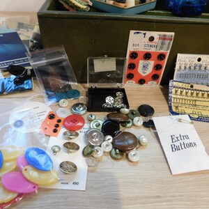 May include: A collection of various buttons in different colors, shapes, and sizes, including black, red, blue, and pearl. Some buttons are on cards, in plastic bags, and in a small black case. A tag reads "Extra Buttons."