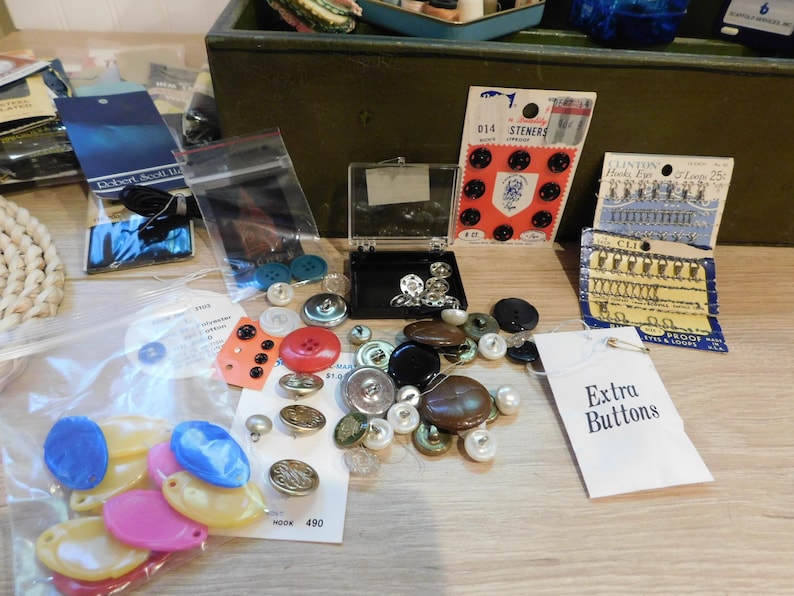 May include: A collection of various buttons in different colors, shapes, and sizes, including black, red, blue, and pearl. Some buttons are on cards, in plastic bags, and in a small black case. A tag reads "Extra Buttons."