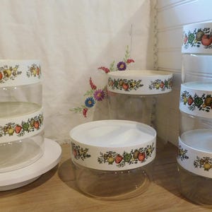 May include: A collection of clear glass storage containers with white lids and decorative bands featuring a colorful fruit and vegetable pattern. The containers are stacked in various configurations, showcasing their design and functionality.