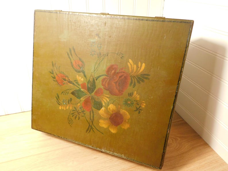 May include: A vintage wooden box with a painted floral design. The flowers are in shades of red, yellow, and orange, with green leaves and stems. The box has a distressed, olive green background and is likely used for storage.
