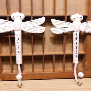 May include: Two white dragonfly-shaped wall hooks with distressed detailing. Each hook features a dragonfly body with wings and a curved hook at the bottom. The hooks are mounted on a wooden background.