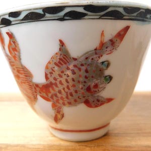May include: White ceramic teacup with a hand-painted design of a red and gray fish. The fish has detailed scales and fins. The rim of the cup has a black and blue leaf pattern. The teacup is a traditional Asian design.