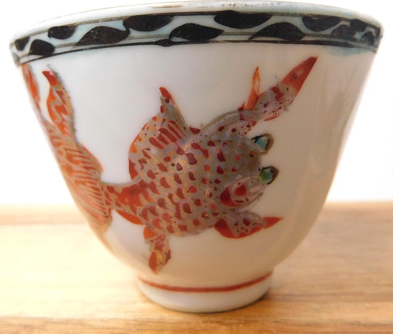 May include: White ceramic teacup with a hand-painted design of a red and gray fish. The fish has detailed scales and fins. The rim of the cup has a black and blue leaf pattern. The teacup is a traditional Asian design.