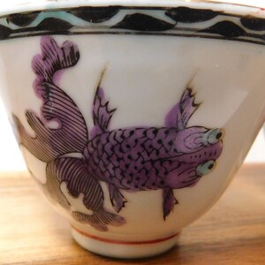 May include: White porcelain teacup with a purple fish design. The fish has detailed scales and fins, with a black and white patterned border at the rim. The cup has a red line near the base.