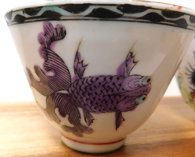 May include: White porcelain teacup with a purple fish design. The fish has detailed scales and fins, with a black and white patterned border at the rim. The cup has a red line near the base.