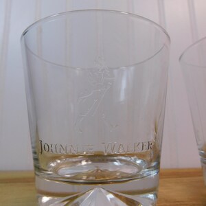 Pode incluir: Copo de vidro transparente gravado com as palavras "JOHNNIE WALKER" e a figura de um homem a andar. O vidro tem uma base espessa e facetada e um topo ligeiramente curvo. O fundo &eacute; uma superf&iacute;cie de madeira clara.
