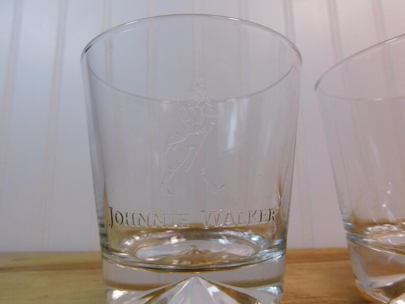 Pode incluir: Copo de vidro transparente gravado com as palavras "JOHNNIE WALKER" e a figura de um homem a andar. O vidro tem uma base espessa e facetada e um topo ligeiramente curvo. O fundo &eacute; uma superf&iacute;cie de madeira clara.