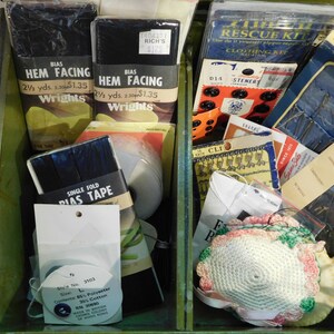 May include: A green wooden box filled with sewing supplies. Includes black hem facing, bias tape, buttons, needles, and a sewing kit. Various packaging with text labels are visible.