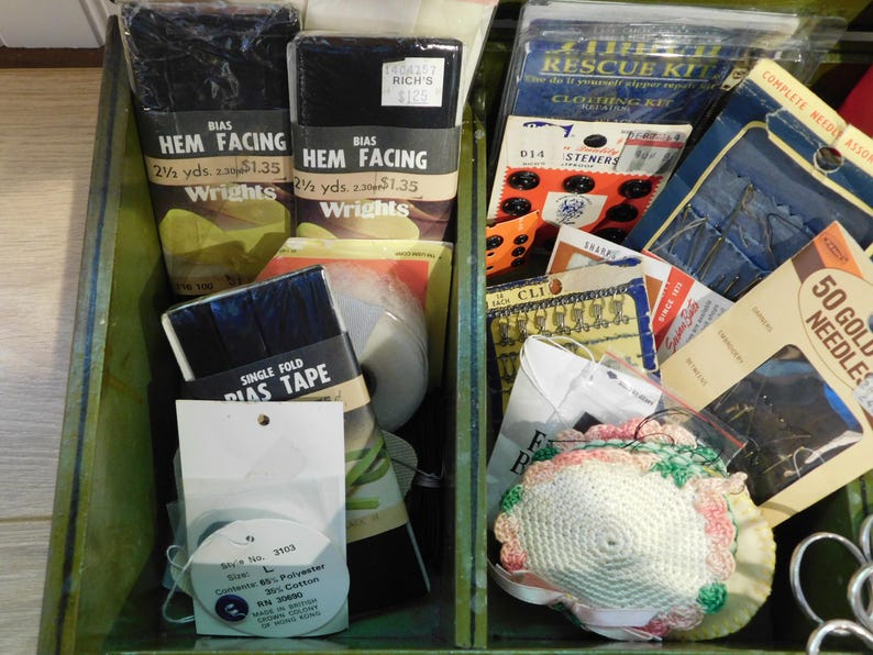 May include: A green wooden box filled with sewing supplies. Includes black hem facing, bias tape, buttons, needles, and a sewing kit. Various packaging with text labels are visible.